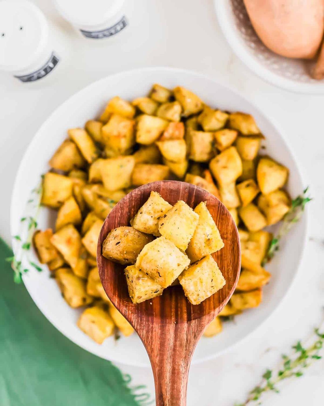 A wooden spoon holds seasoned, roasted cubed sweet potatoes over a white plate filled with more of the same. Fresh herbs and a raw sweet potato are visible in the background.