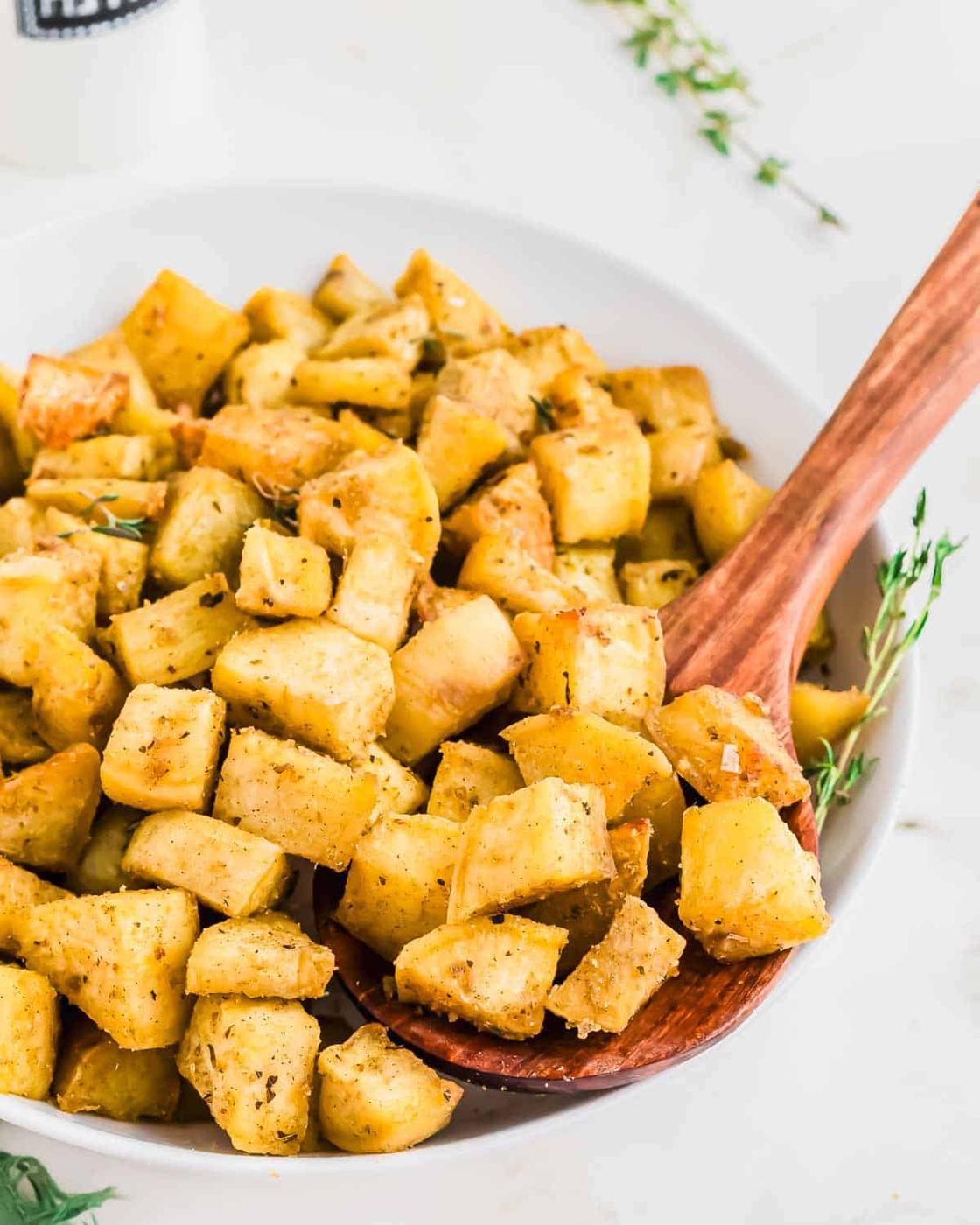 A white bowl filled with seasoned, roasted Japanese white sweet potatoes, with a wooden spoon scooping some out. Fresh herbs are scattered around.