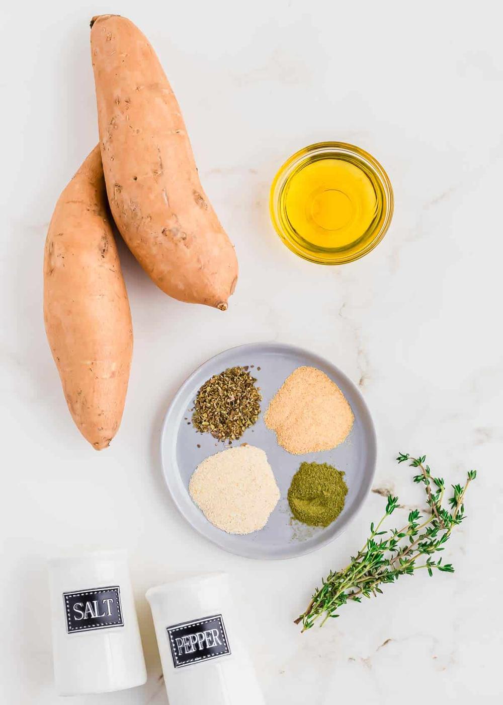 Two white sweet potatoes, a bowl of olive oil, a plate with four spices, salt and pepper shakers, and a sprig of fresh herbs are arranged on a white surface.