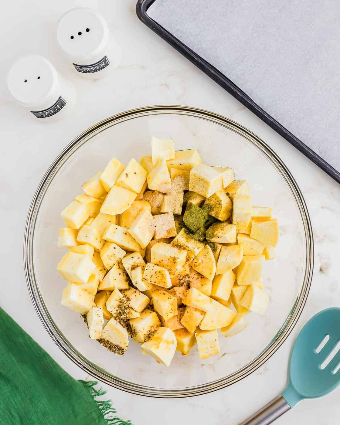 A glass bowl containing cubed potatoes seasoned with various spices, a baking tray lined with parchment paper, and a green spatula on a white surface. Two spice shakers are nearby.