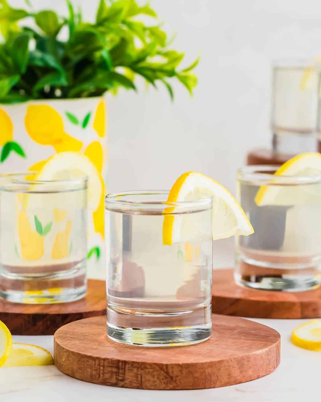 Three glasses of white tea shots, each garnished with a lemon slice, are placed on wooden coasters. A potted plant with a lemon-themed pot is in the background.