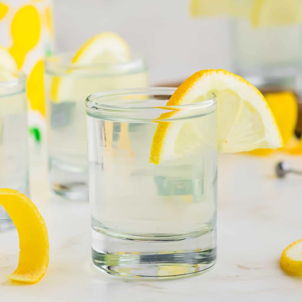 A clear drink in a short, transparent glass garnished with a lemon wedge. Other similar glasses with lemon slices are in the background, along with lemon peels on the surface.