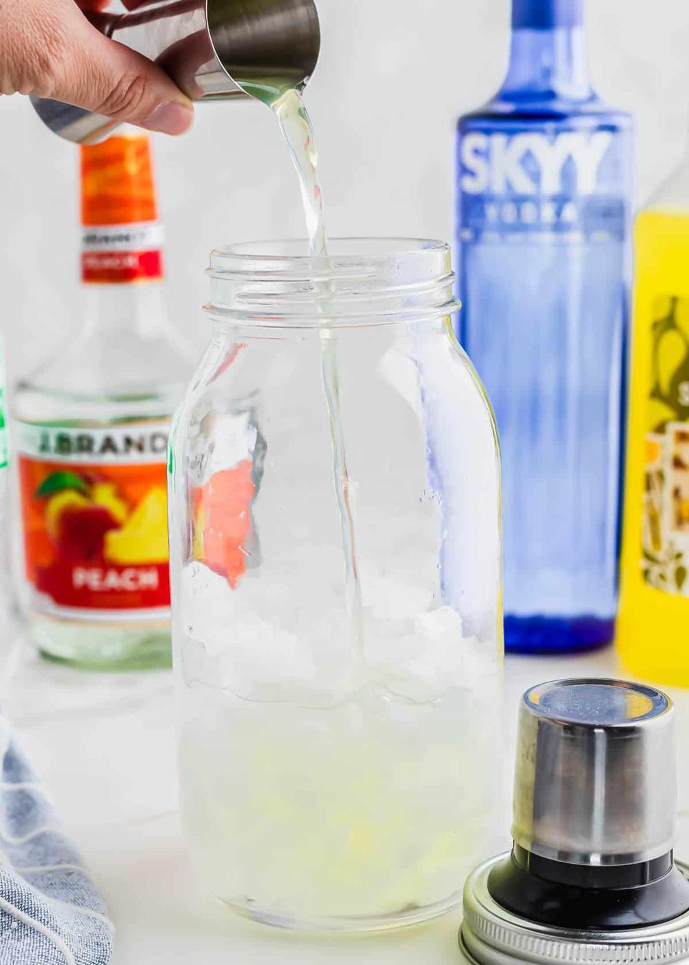 A hand pours a liquid from a shot measuring cup into a mason jar filled with ice. Bottles of alcohol, including vodka and peach schnapps, are visible in the background. A cocktail shaker is on the table.
