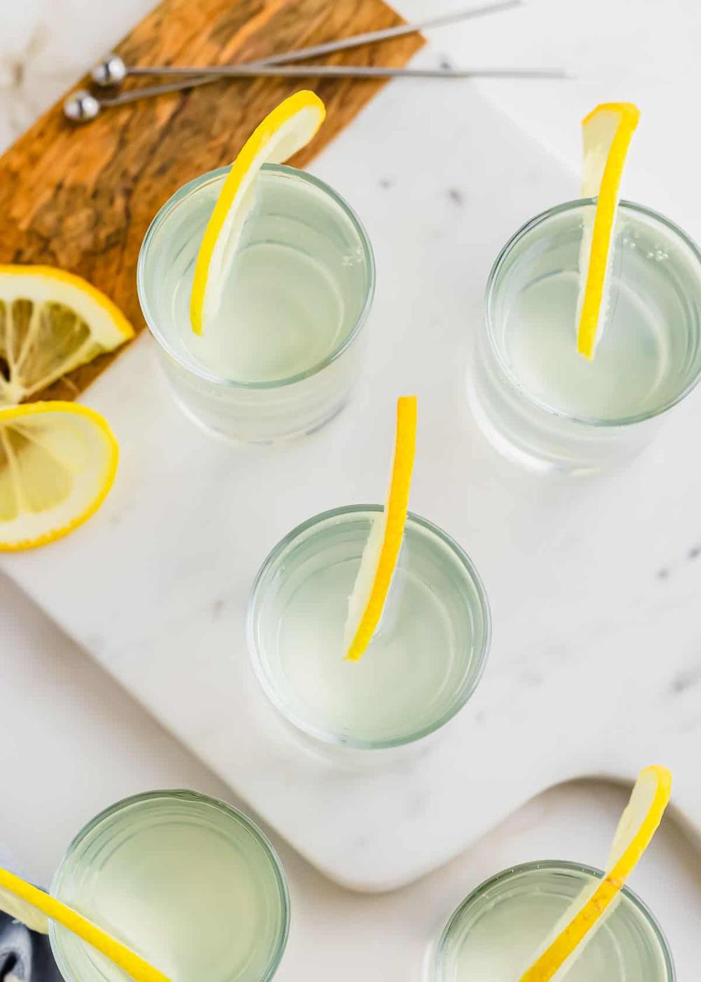 Four white tea shots, garnished with lemon wedges, are arranged on a white marble surface with additional lemon slices and metal serving tongs nearby.