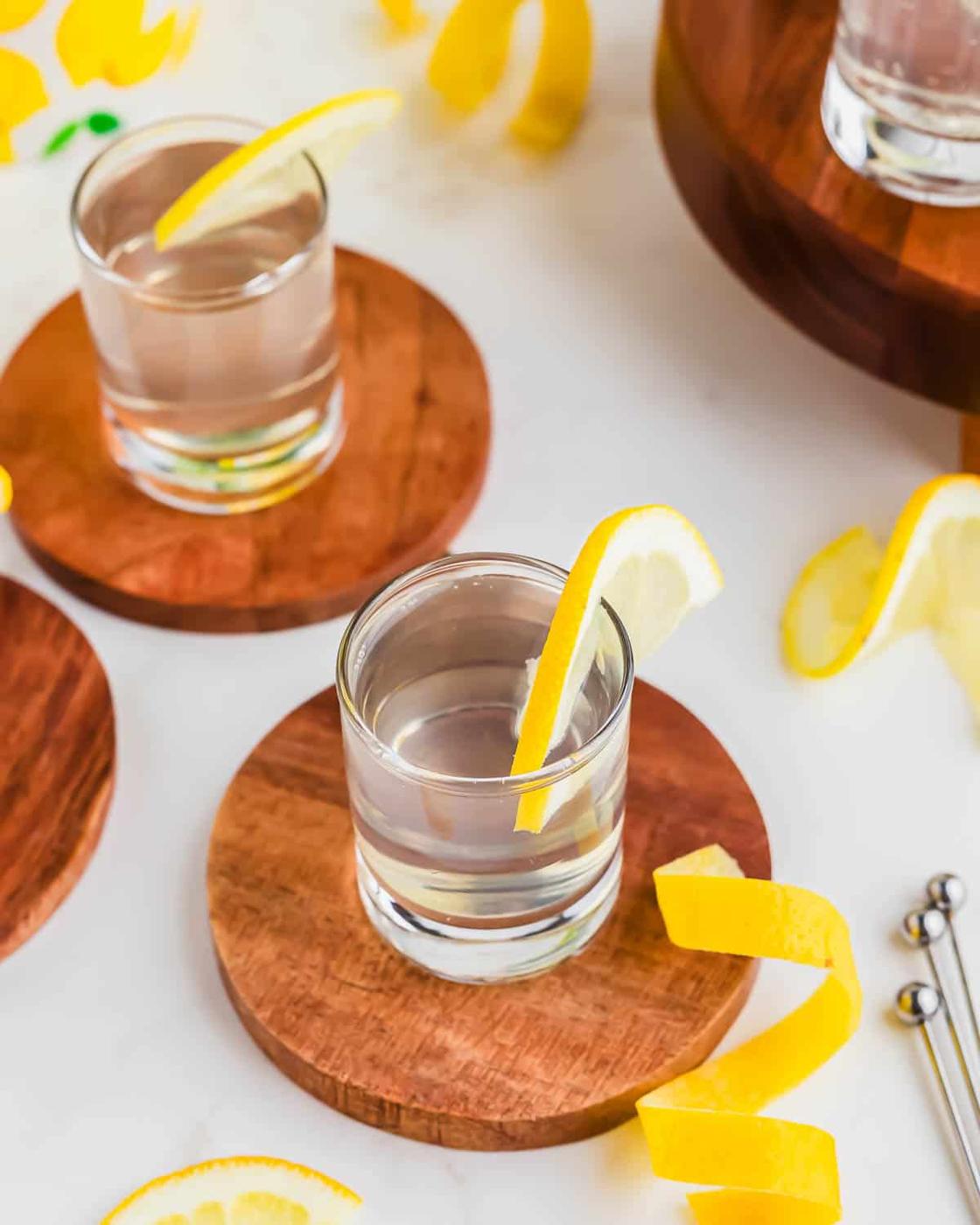 A white tea shot glass with a lemon slice on the rim sits on a wooden coaster surrounded by lemon peels and another identical drink.