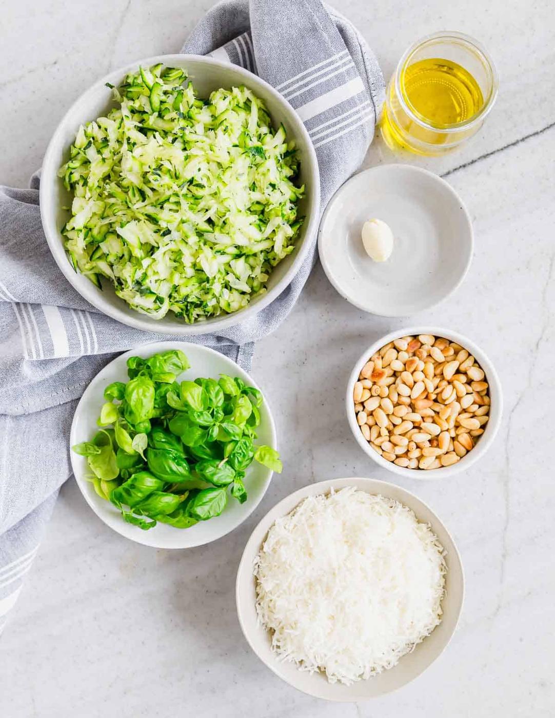 Ingredients for pesto laid out on a marble surface: shredded zucchini, a glass of oil, garlic clove, pine nuts, fresh basil, grated cheese, and a gray-striped napkin.