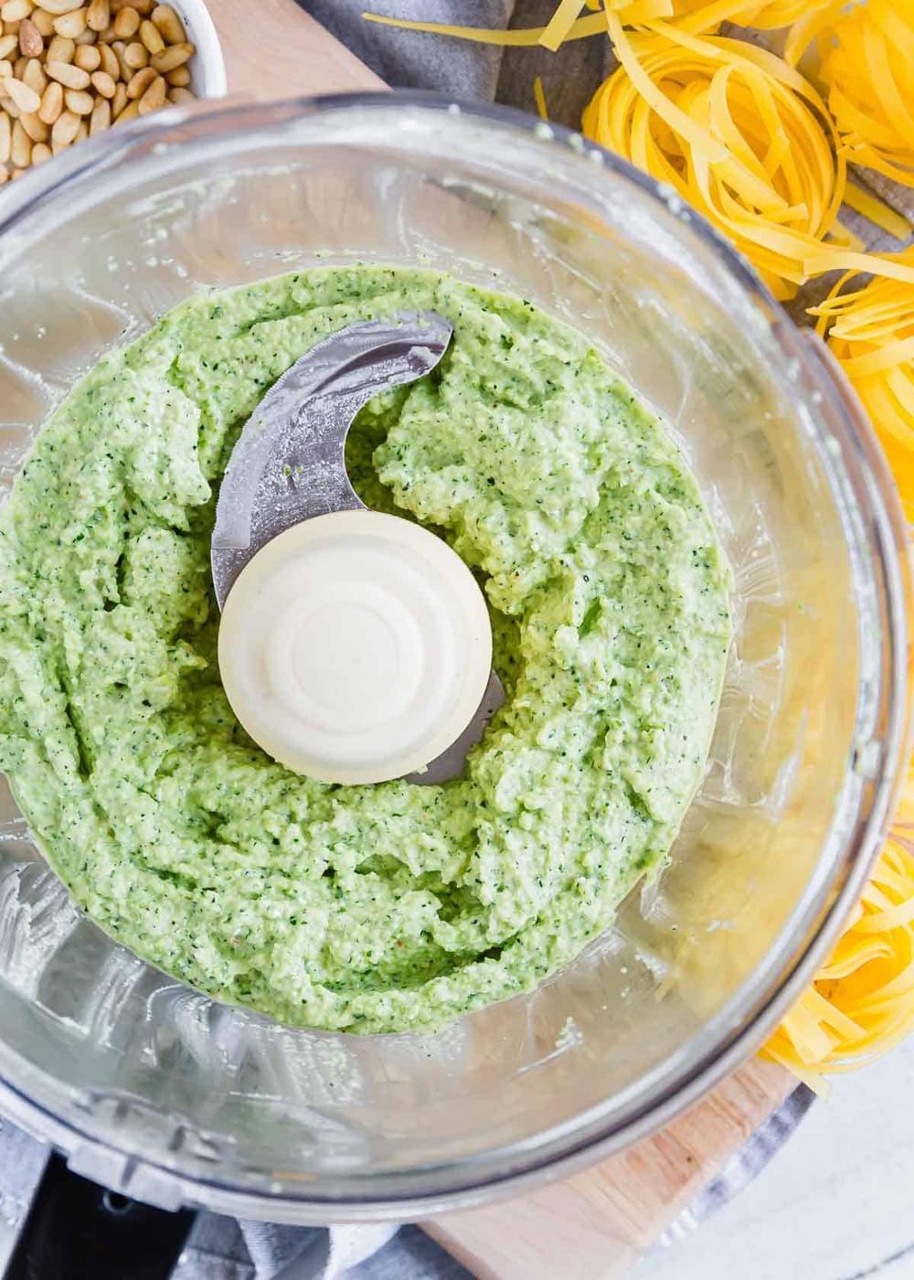A food processor containing green zucchini pesto with a visible blade is placed on a wooden surface next to uncooked yellow pasta and a bowl of pine nuts.