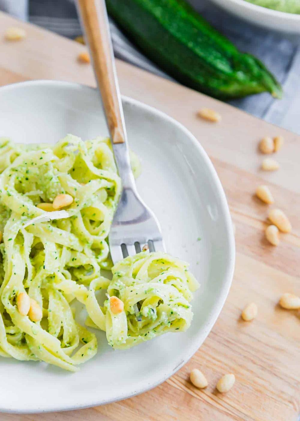 A fork with tangled zucchini pesto pasta topped with a few pine nuts sits on a white plate, next to a whole zucchini in the background.