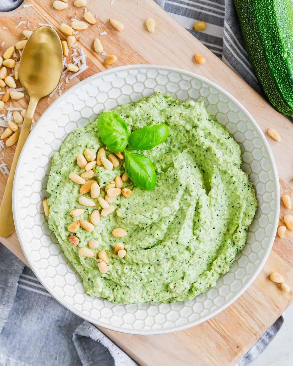 A bowl of green pesto, garnished with basil leaves and pine nuts, placed on a wooden board. A gold spoon and scattered pine nuts are beside the bowl. A zucchini is nearby.