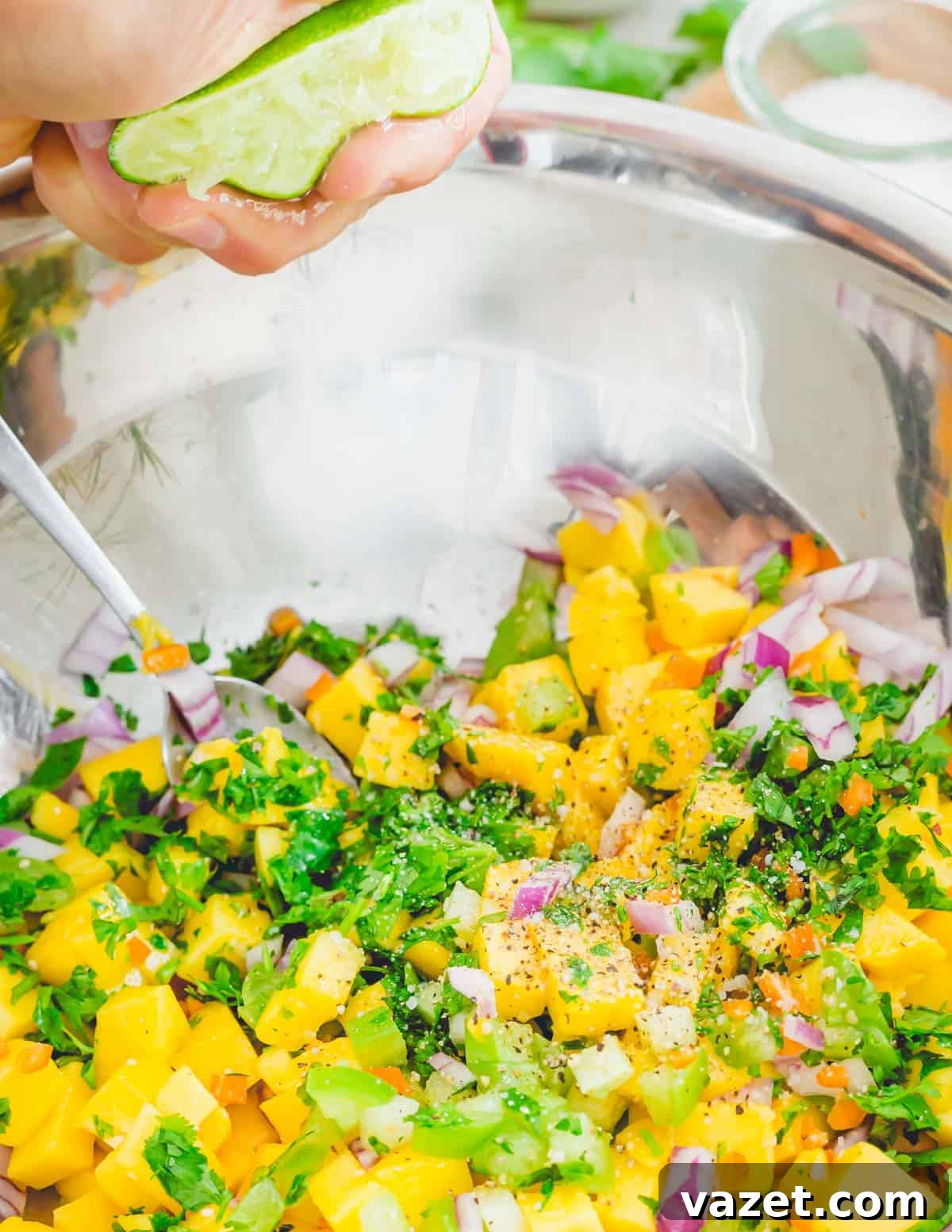 A hand squeezing fresh lime juice into a bowl filled with diced mango, cilantro, tomatillos, red onion, and habanero peppers, preparing to mix the salsa.
