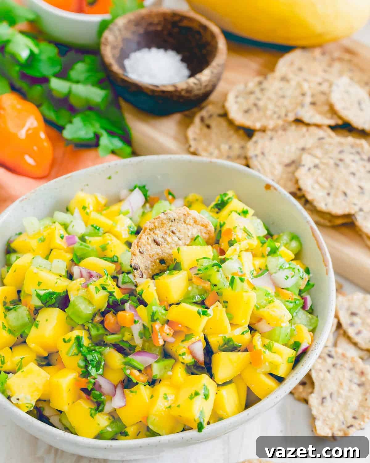 A cracker partially submerged in a bowl of vibrant mango salsa, with clear views of diced habanero peppers, cilantro, red onion, tomatillos, and the glistening lime juice.