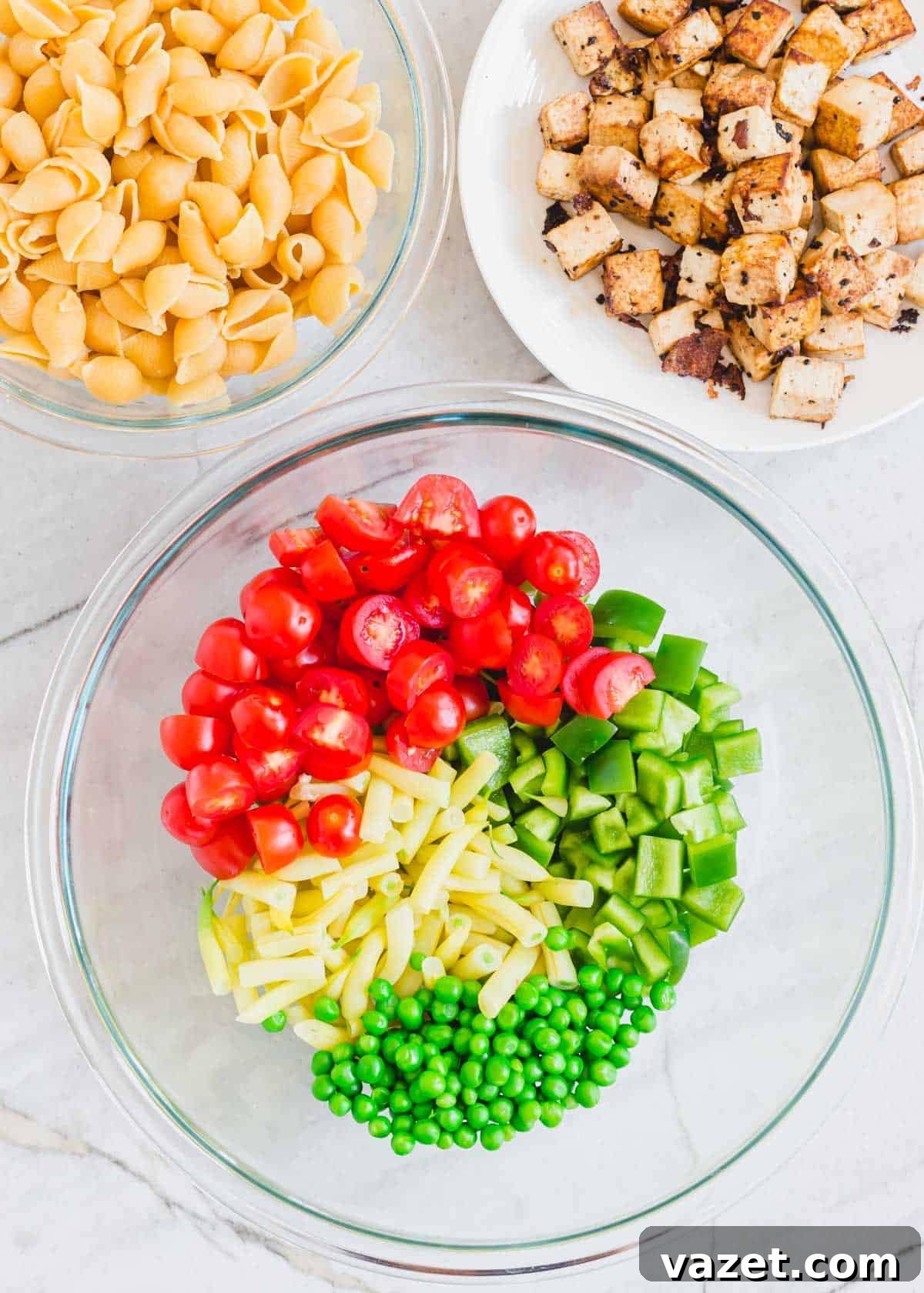 Fresh Ingredients for a Healthy Tofu Pasta Salad Ingredients for Tofu Pasta Salad: cooked shell pasta, crispy tofu, cherry tomatoes, green bell peppers, peas, and yellow wax beans displayed in glass bowls and on a plate.