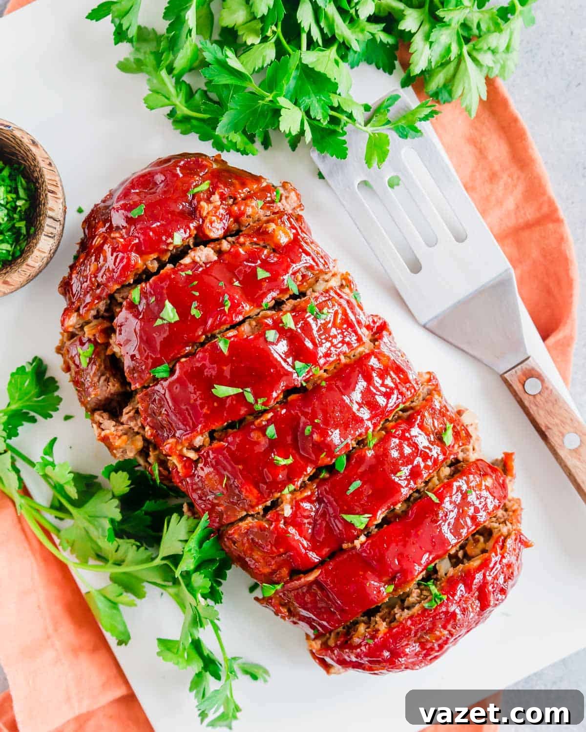 Cooked meatloaf sliced on a white plate with fresh parsley and a serving spatula on the side. A perfectly cooked meatloaf, glistening with glaze, presented on a white plate with a serving spatula, ready to be enjoyed, garnished with fresh parsley.
