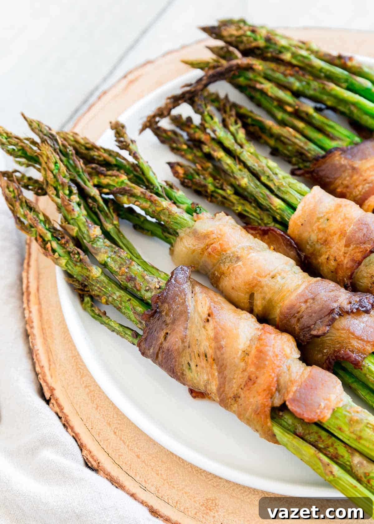 Air Fried Bacon Wrapped Asparagus Perfection 4 Air fryer bacon wrapped asparagus bundles on a white plate with wooden surface beneath.