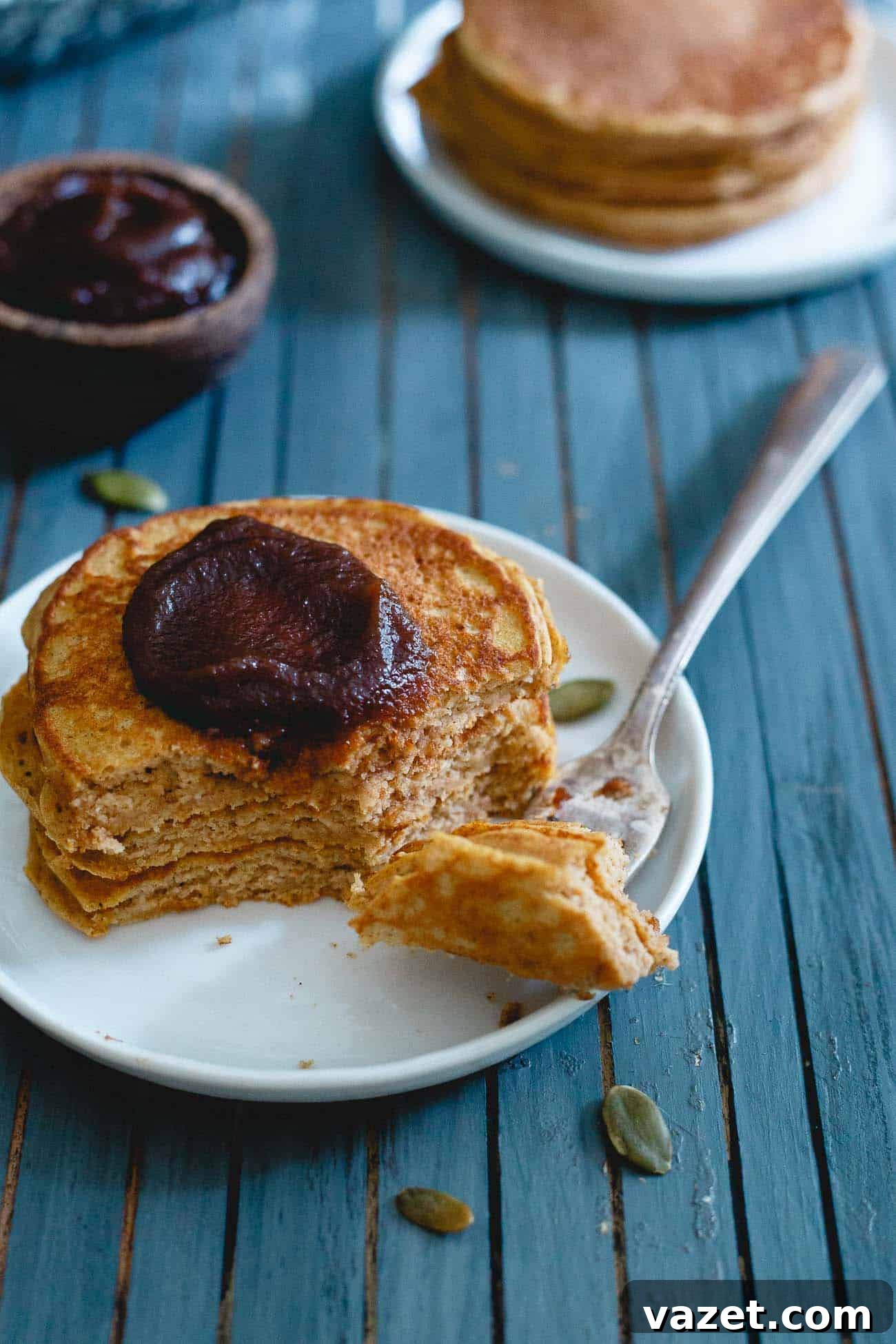 A close-up of a stack of fluffy pumpkin protein pancakes topped with butter and a generous pour of maple syrup, against a fall backdrop.
