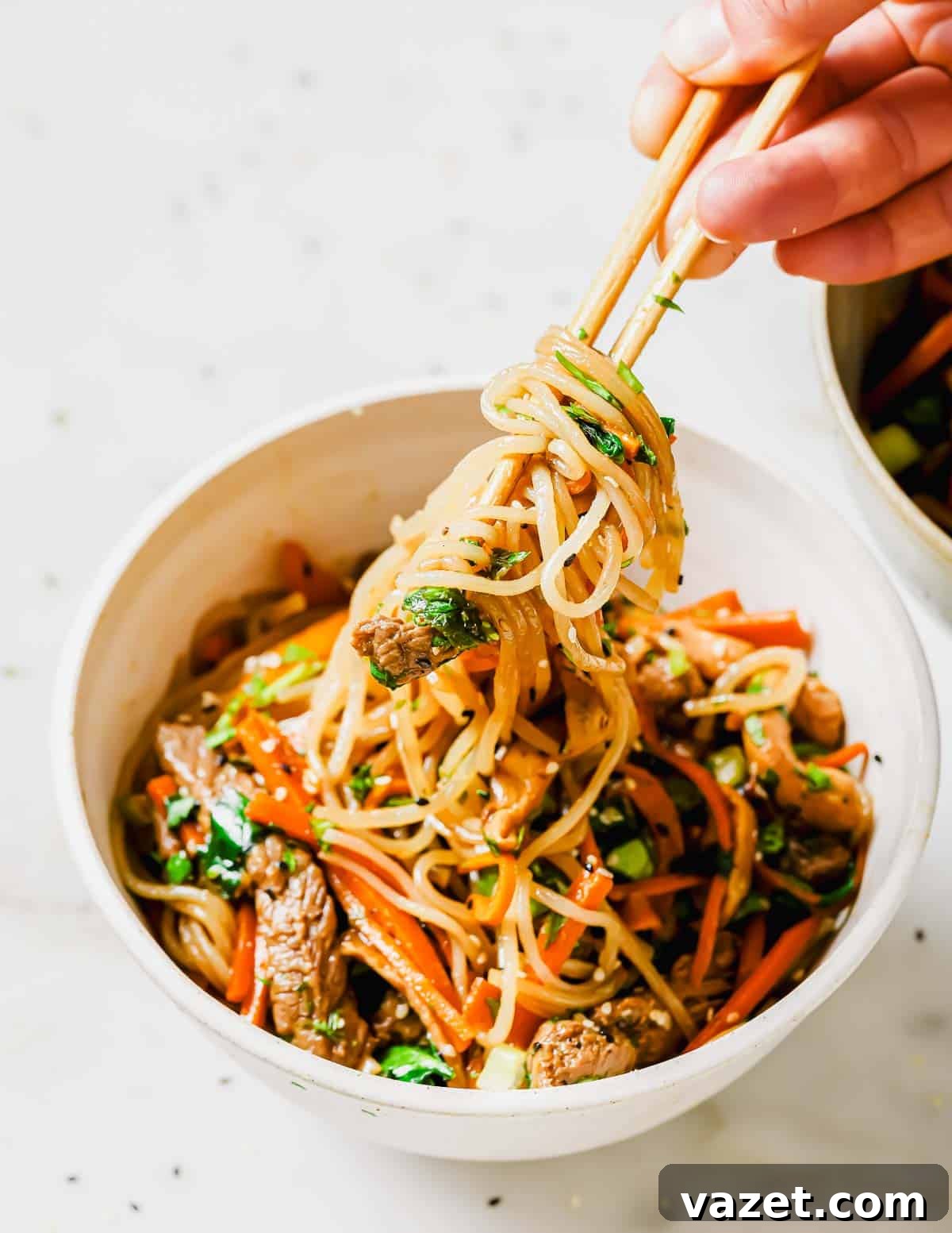A person holding chopsticks over a generously filled bowl of Korean sweet potato glass noodles with beef and vegetables, ready to enjoy.