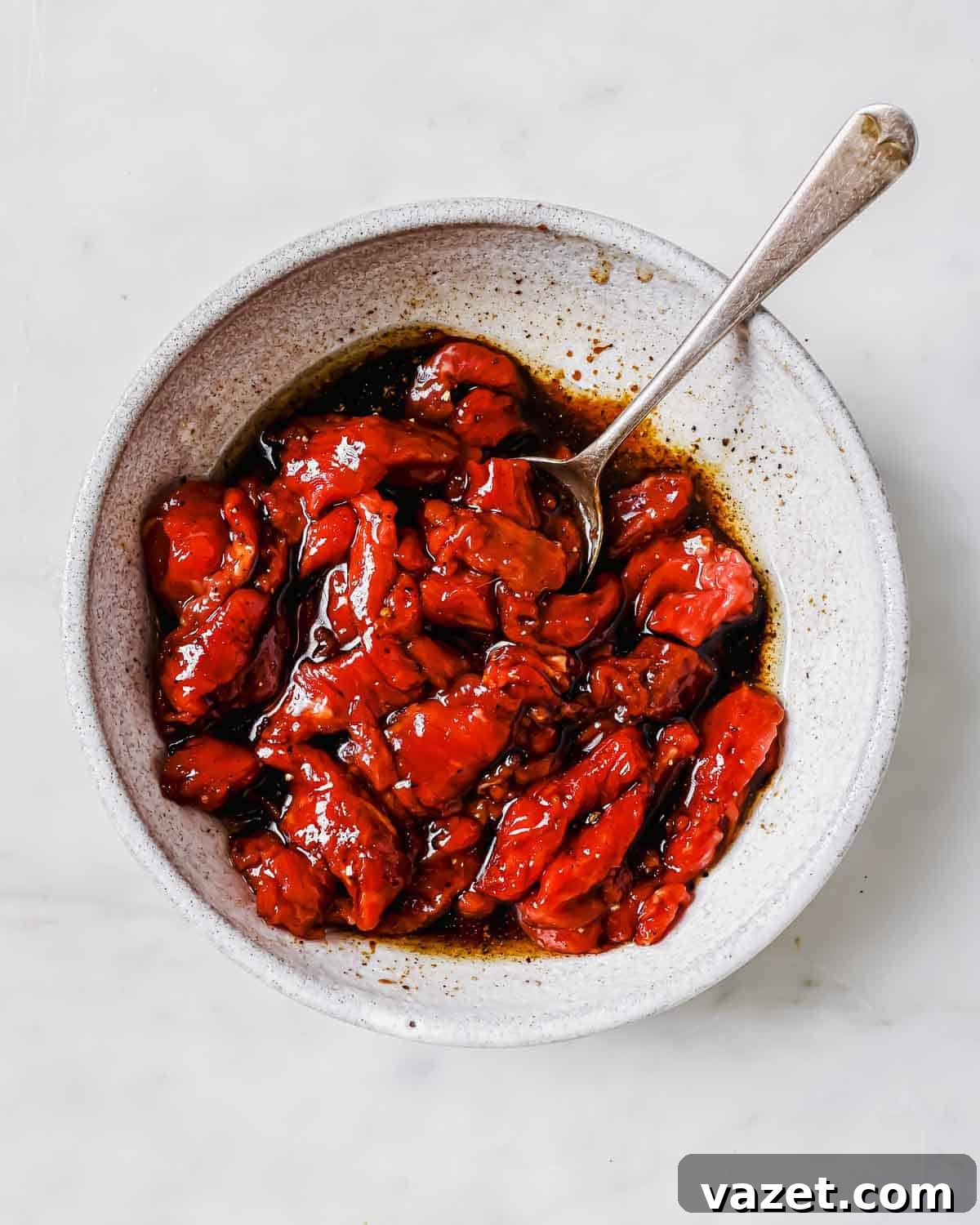 Thin strips of beef marinating in a rich, dark sauce in a white bowl, accompanied by a spoon for stirring.