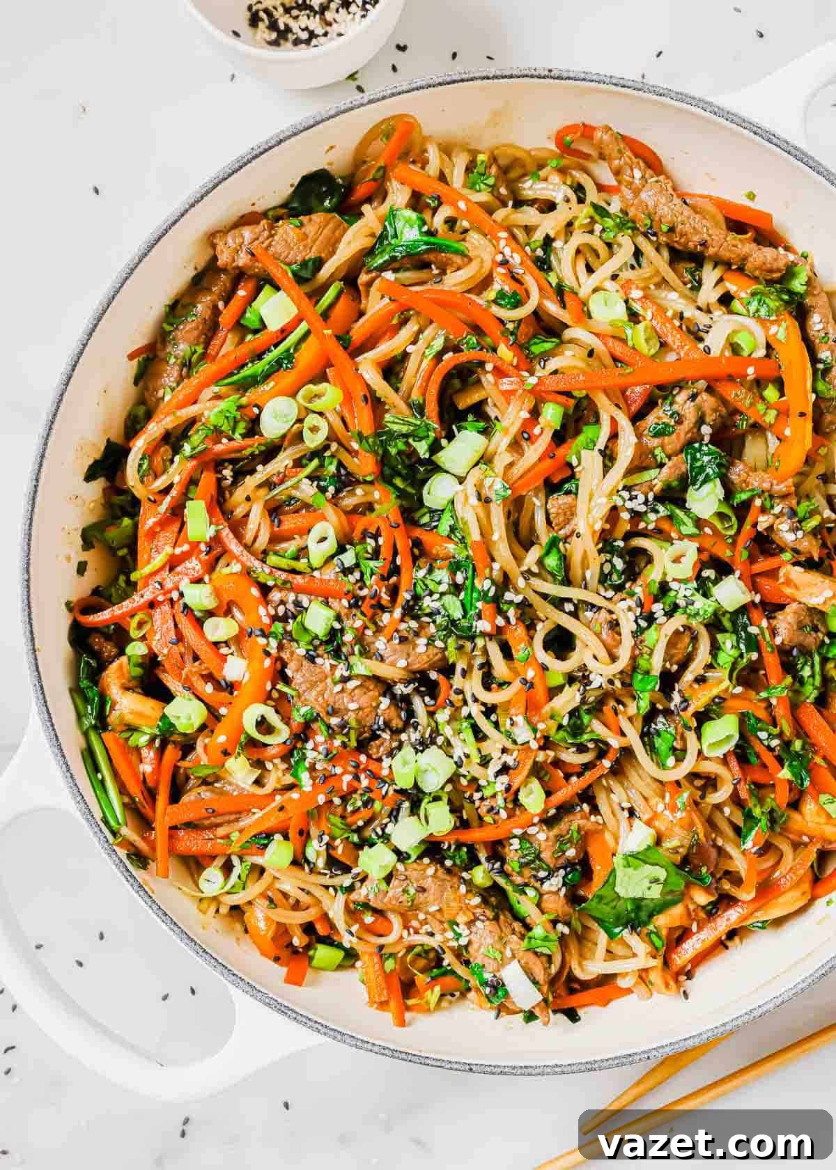 A skillet filled with vibrant Korean sweet potato glass noodles, tender beef, and colorful vegetables, with a pair of chopsticks poised to serve.