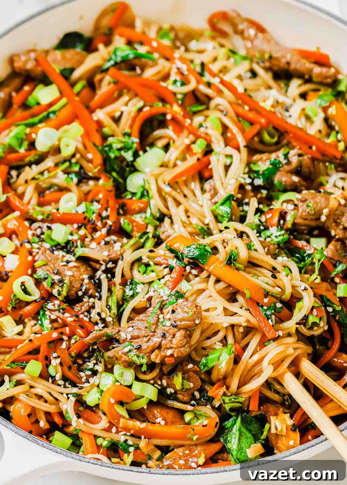 A close-up shot of Korean Japchae, featuring glistening sweet potato glass noodles, beef, and colorful vegetables, in a pan with chopsticks ready to serve.