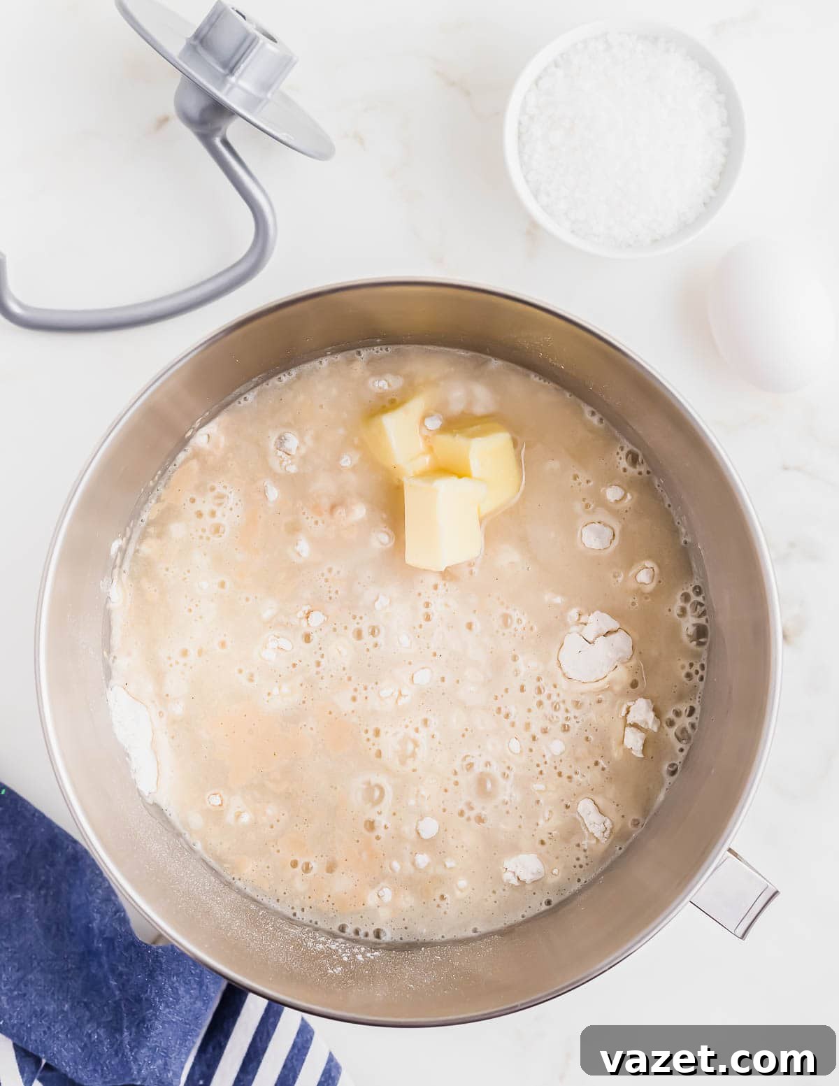 Cloud-Soft Gluten-Free Pretzels 4 A mixing bowl with flour, water, butter, and other ingredients. Nearby are a mixer attachment, a small bowl of coarse salt, an egg, and a blue cloth on a white countertop.