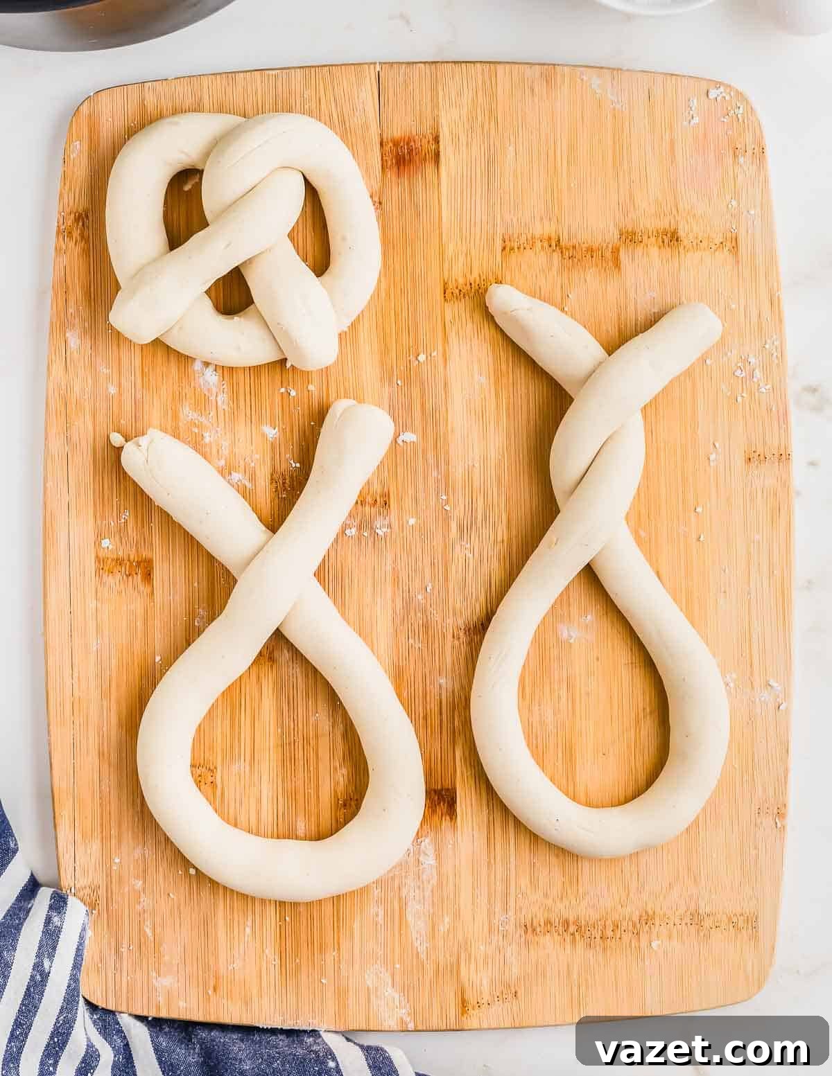 Cloud-Soft Gluten-Free Pretzels 8 Three pretzel dough shapes placed on a wooden cutting board. The top left one resembles a traditional pretzel shape, while the other two are partially twisted into an elongated form.