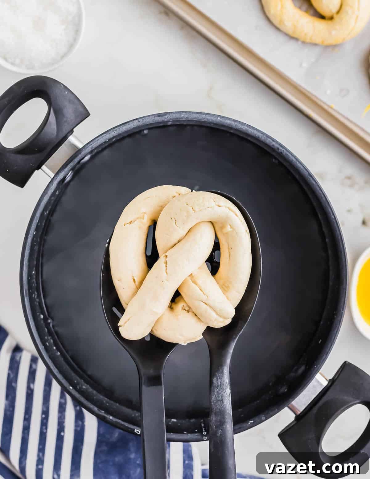 Cloud-Soft Gluten-Free Pretzels 9 A partially baked pretzel on a slotted spoon hovers over a pot, ready for boiling. Other kitchen utensils and ingredients are visible in the background.