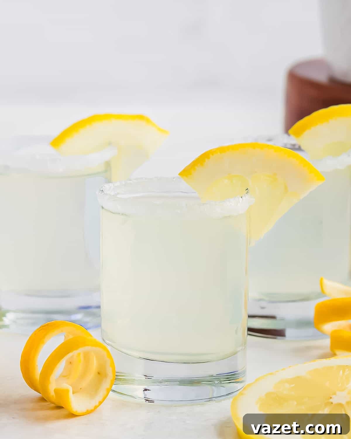 Zesty Lemon Drop Shot 2 Three glasses of lemon drop shots, garnished with lemon slices on the rims. The glasses have sugared edges and are placed on a white surface, alongside lemon slices and twists. A blurred background with a wooden element is visible.