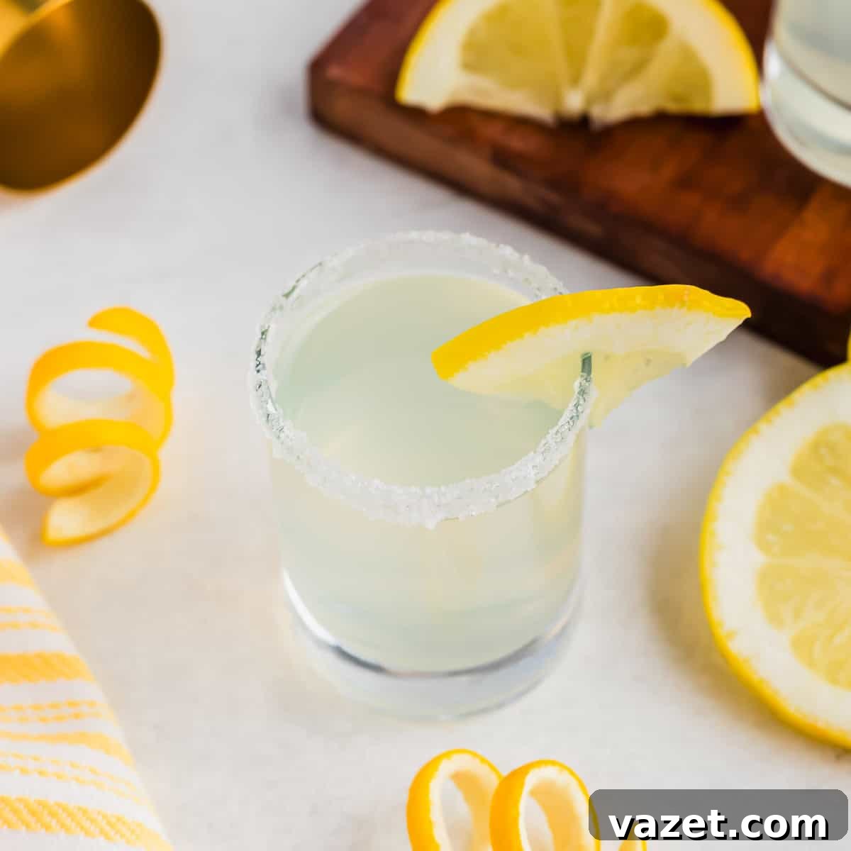 Zesty Lemon Drop Shot 11 A glass of lemon drop shot cocktail with a sugared rim and lemon wedge garnish sits on a white surface. Nearby are lemon slices and peels, a yellow-striped cloth, and a wooden board with more lemon slices.