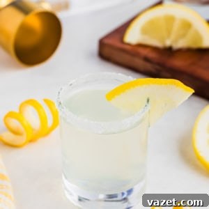 Zesty Lemon Drop Shot 12 A salt-rimmed glass with a lemon wedge garnish contains a lemon drop shot. Curled lemon peels and sliced lemons are scattered around on the white surface. A wooden board, golden jigger, and cloth are visible in the background.