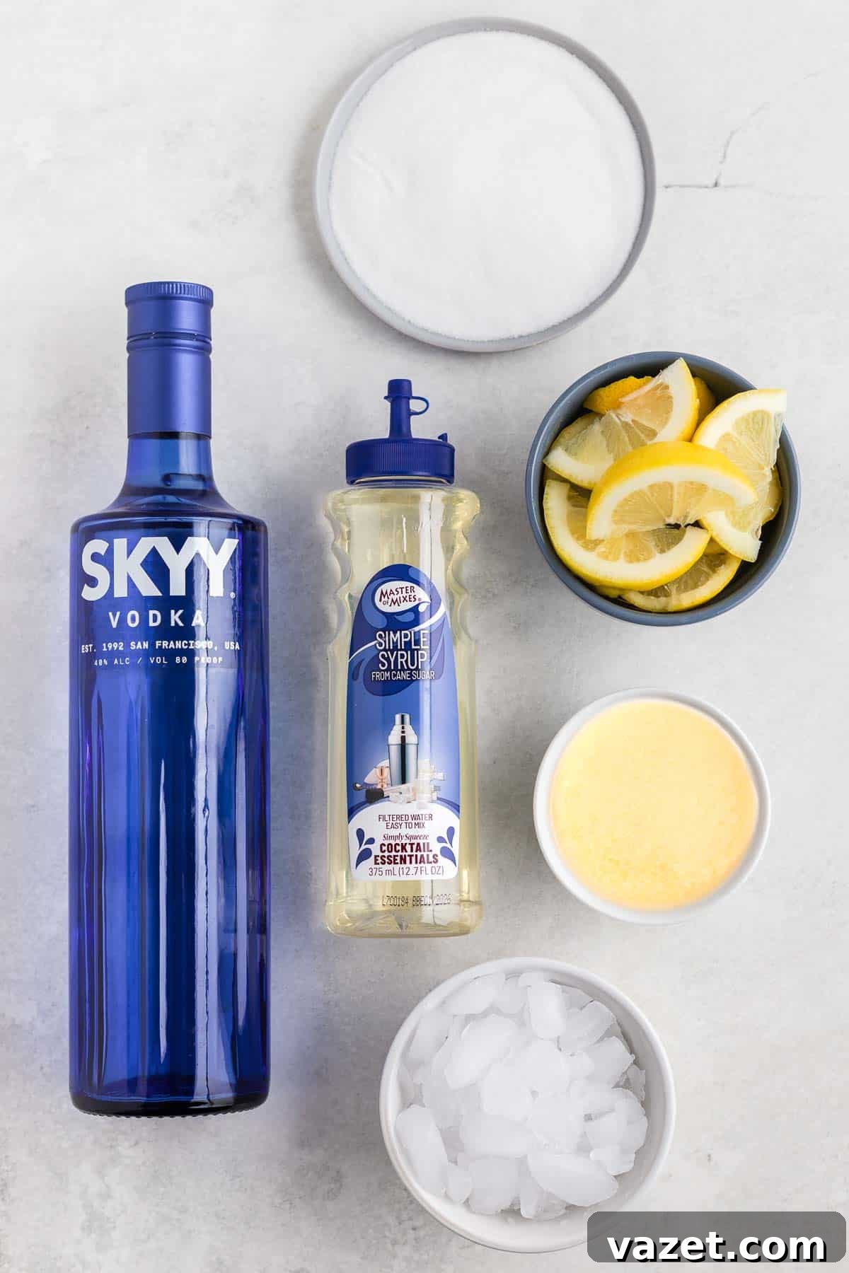 Zesty Lemon Drop Shot 3 Overhead view of cocktail ingredients: a blue bottle of Skyy vodka, a bottle of simple syrup, a bowl of sugar, a bowl of lemon slices, a bowl of lemon juice, and a bowl of ice on a light surface.