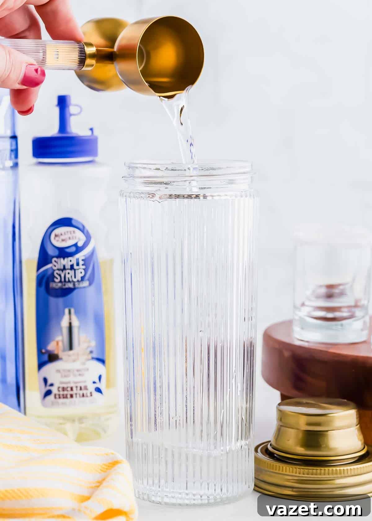Zesty Lemon Drop Shot 5 A hand pouring a liquid from a brass measuring cup into a tall, ribbed glass. A bottle of simple syrup and other glassware is visible in the background. A gold jar lid and a yellow cloth are on the side.