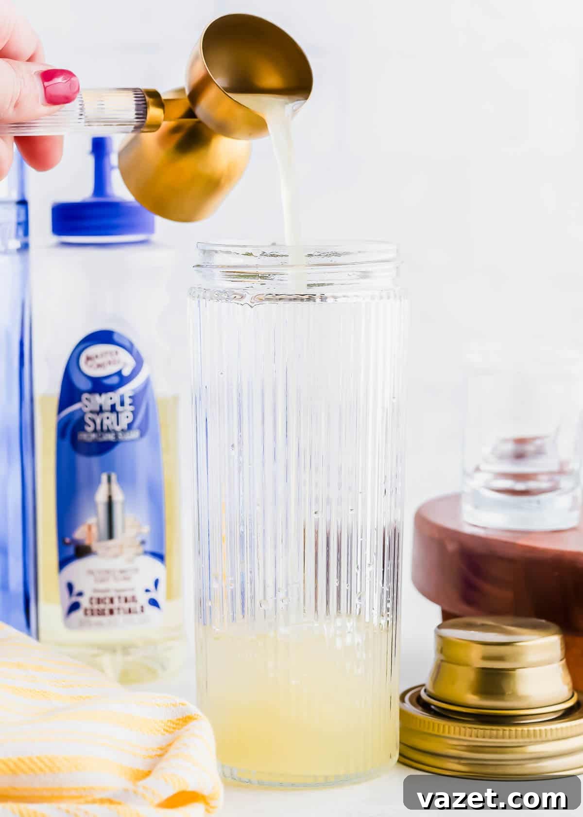Zesty Lemon Drop Shot 6 A hand pours liquid from a brass measuring cup into a tall, ridged glass. Behind the glass, there's a bottle labeled