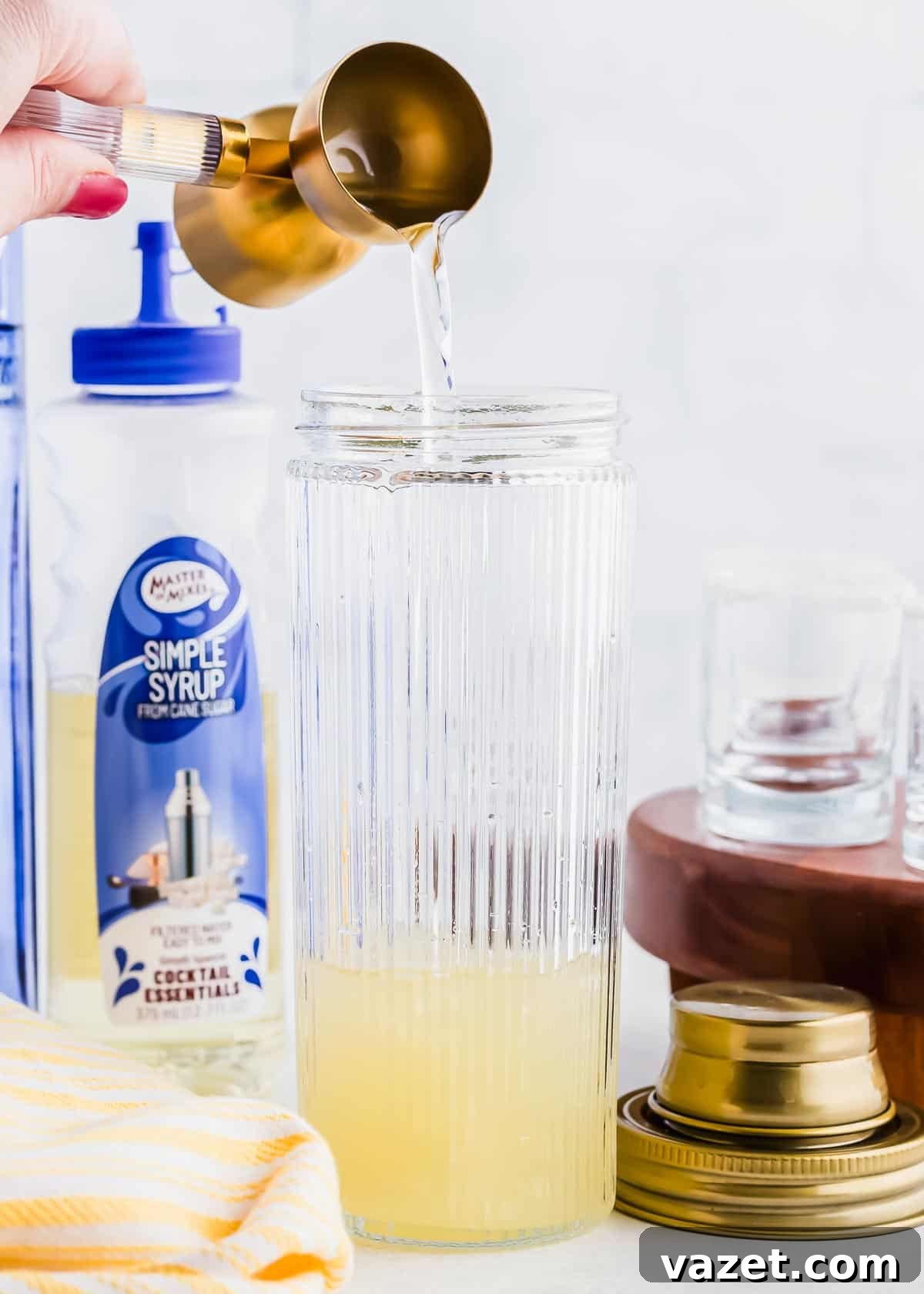 Zesty Lemon Drop Shot 7 A hand pours liquid from a brass measuring cup into a glass cocktail shaker with grooves. Nearby, there's a bottle of simple syrup and stacked shot glasses. A shaker lid is on the surface next to a yellow-striped towel.