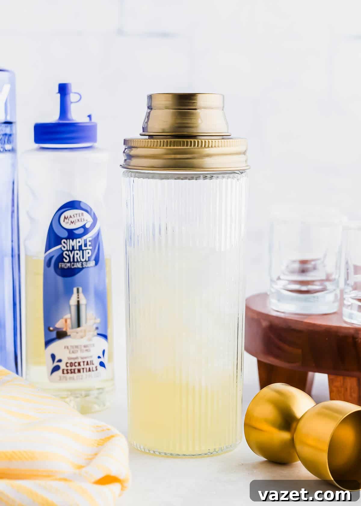 Zesty Lemon Drop Shot 8 A ribbed cocktail shaker filled with liquid stands on a counter, surrounded by a bottle of simple syrup, stacked glass cups on a wooden stand, a gold jigger, and a yellow striped cloth.