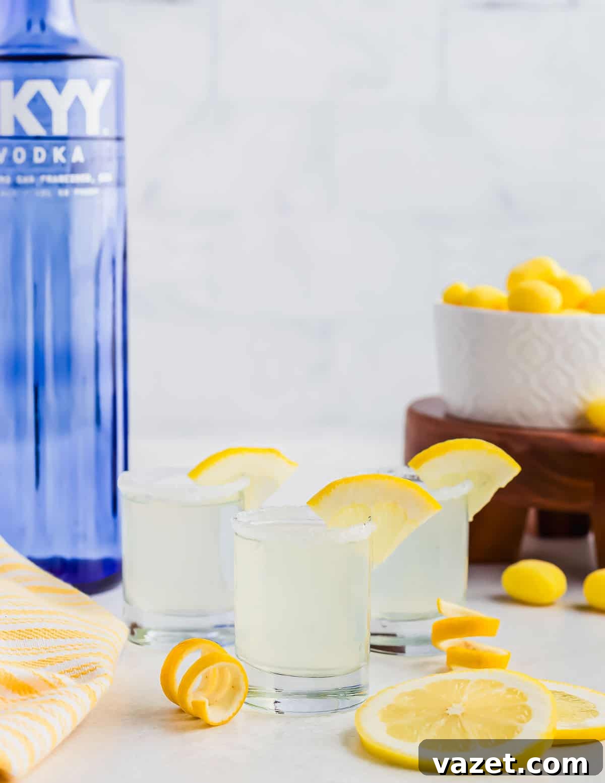 Zesty Lemon Drop Shot 9 Three lemon drop cocktails in shot glasses, garnished with lemon slices, sit on a white surface. A blue vodka bottle and a bowl of yellow candies are in the background. Lemon wedges are scattered around, along with a yellow striped towel.