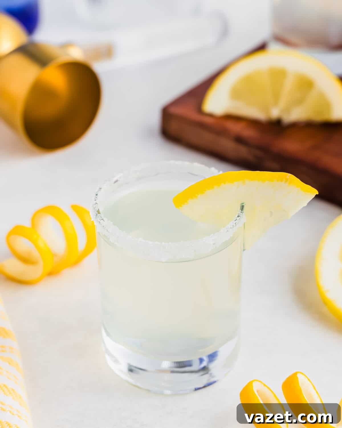 Zesty Lemon Drop Shot 10 A salt-rimmed glass with a lemon wedge garnish contains a lemon drop shot. Curled lemon peels and sliced lemons are scattered around on the white surface. A wooden board, golden jigger, and cloth are visible in the background.