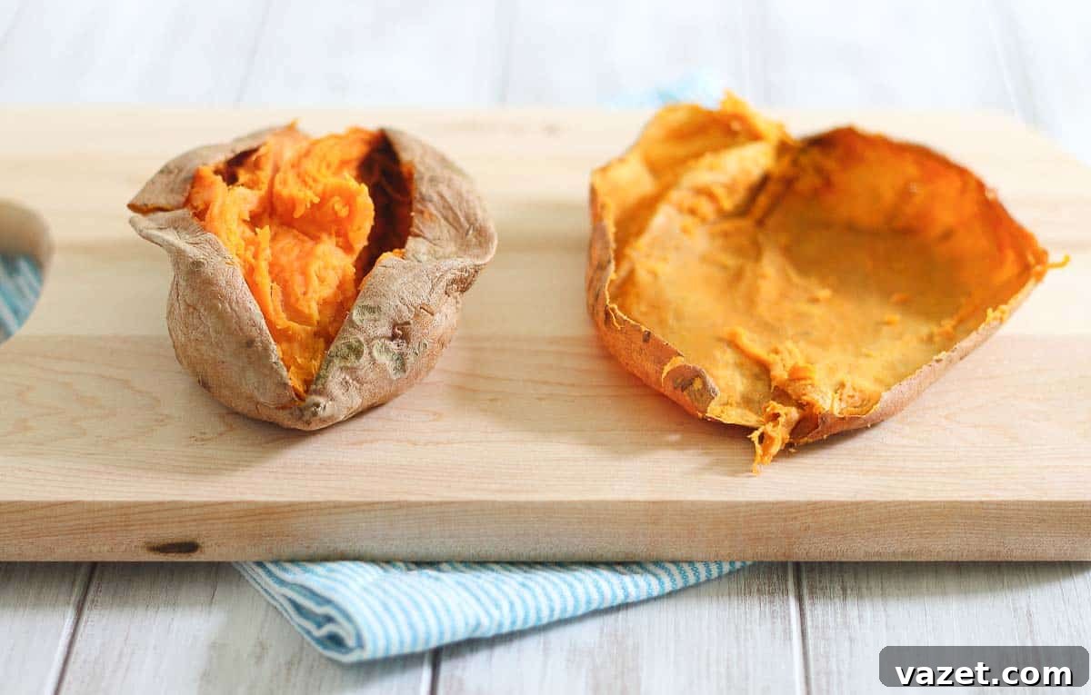 A split baked sweet potato, with the flesh scooped out, lies on a wooden cutting board with a blue-striped cloth underneath.