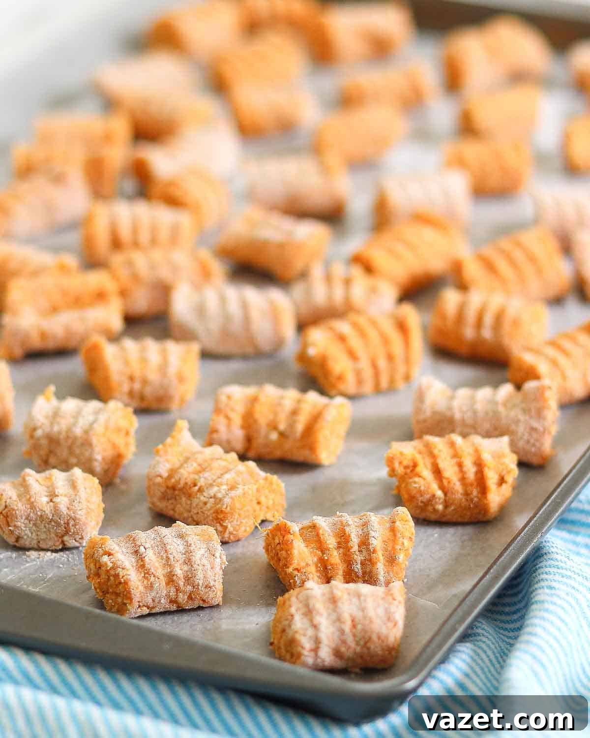A baking tray filled with uncooked homemade sweet potato gnocchi, lightly dusted with flour. The gnocchi pieces are evenly spaced and have a ridged texture. The tray is on a striped blue cloth.