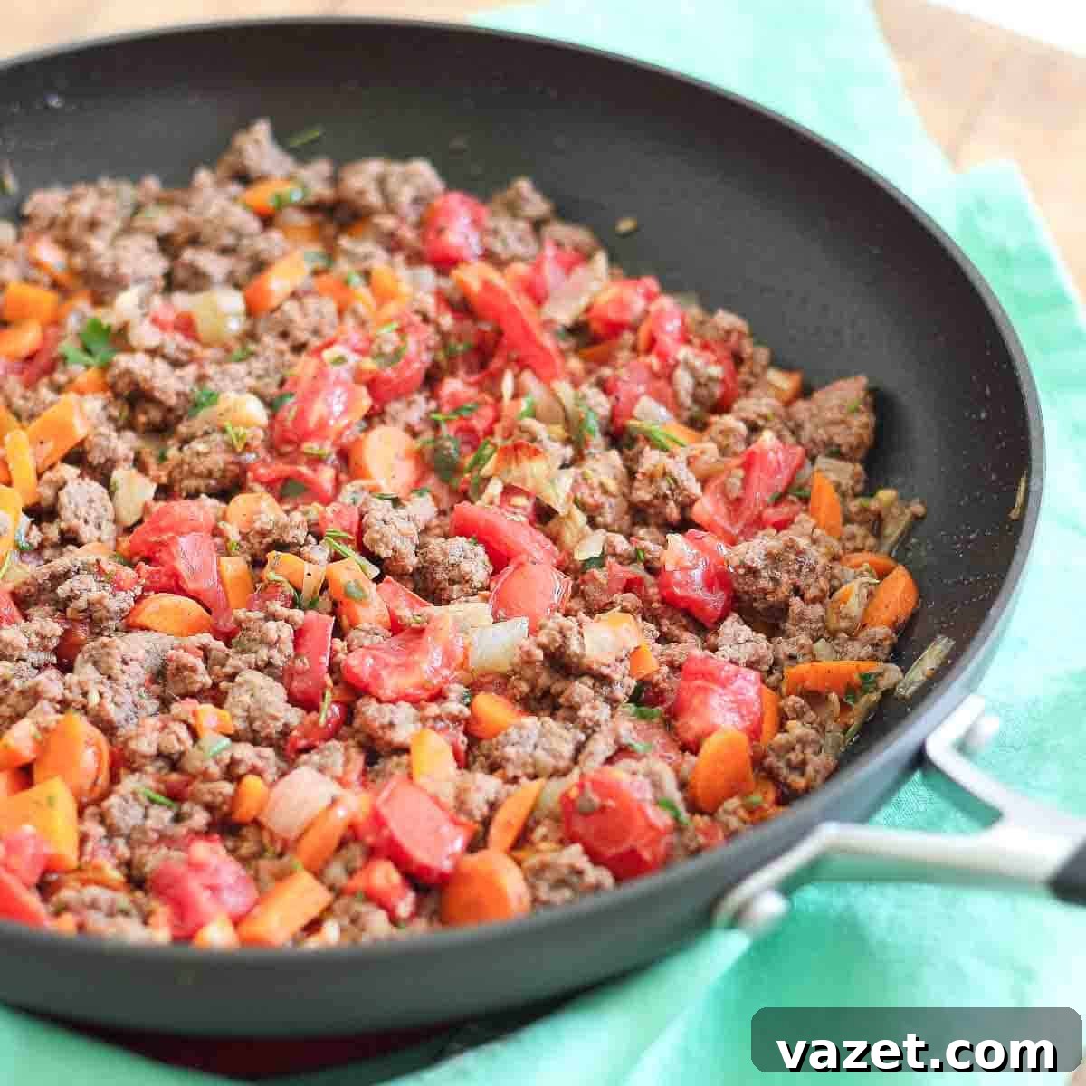 A skillet filled with cooked ground meat, diced tomatoes, carrots, and onions on a green napkin.