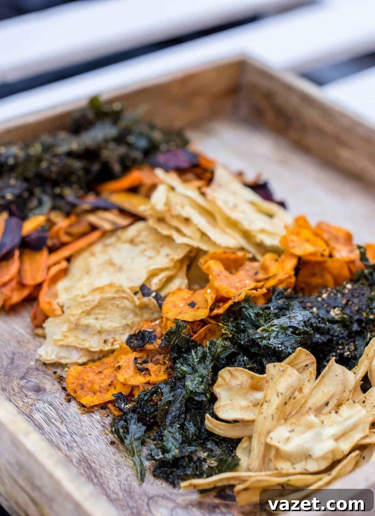 A wooden tray filled with an assortment of dehydrated vegetable chips, including kale, carrots, and possibly sweet potato, arranged in colorful, uneven piles.