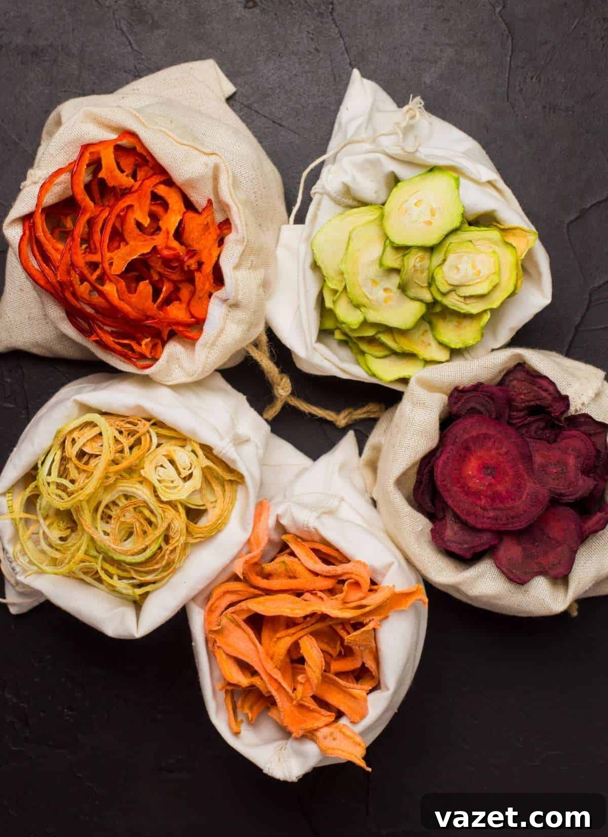 Five small cloth bags filled with colorful dried vegetable chips—orange carrot, green zucchini, yellow squash, purple beet, and orange sweet potato—arranged in a circle on a dark textured surface.