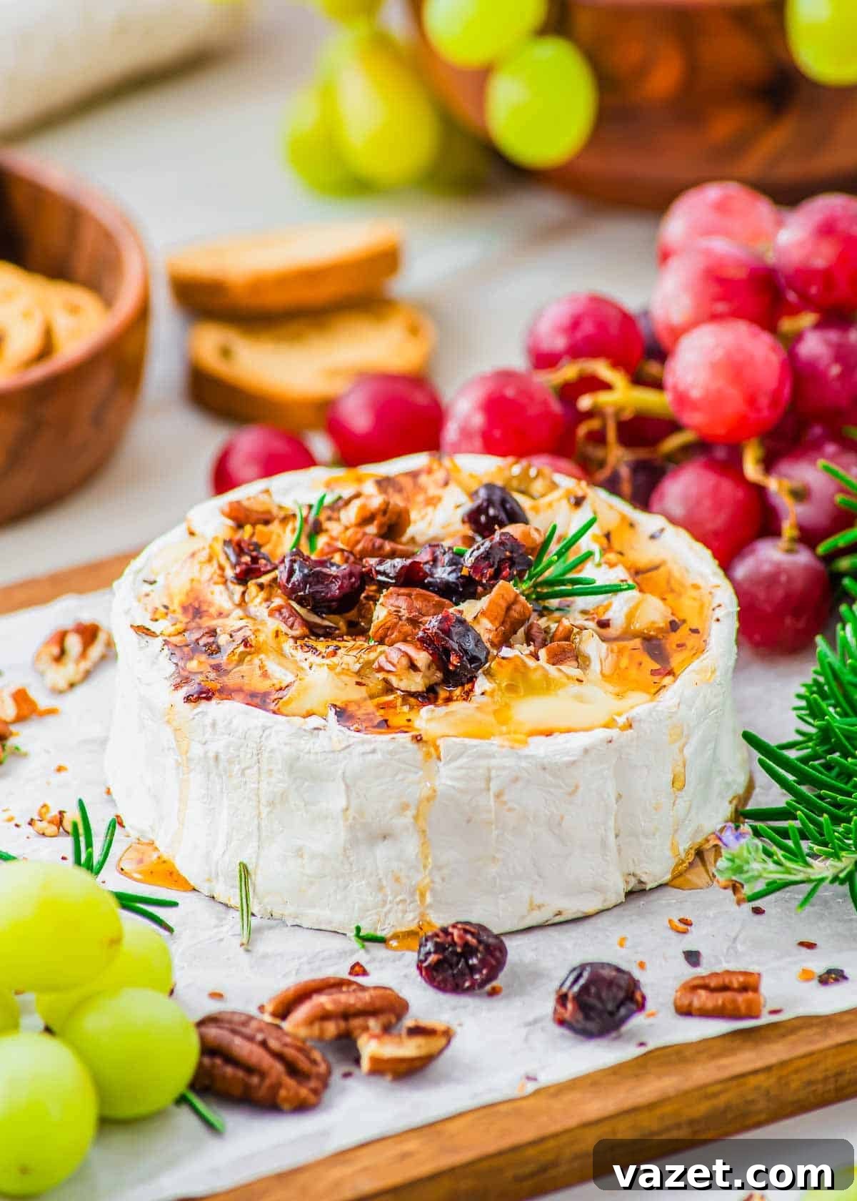 Honey-Kissed Baked Brie 2 A wheel of honey baked brie topped with chopped pecans, dried cranberries and rosemary sits on parchment paper, surrounded by green and red grapes, crackers, and fresh rosemary.
