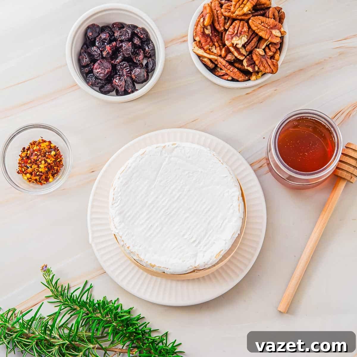 Honey-Kissed Baked Brie 4 A round wheel of brie cheese on a white plate, surrounded by bowls of dried cranberries, pecans, crushed red pepper, a jar of honey with a dipper, and a sprig of fresh rosemary on a light surface.