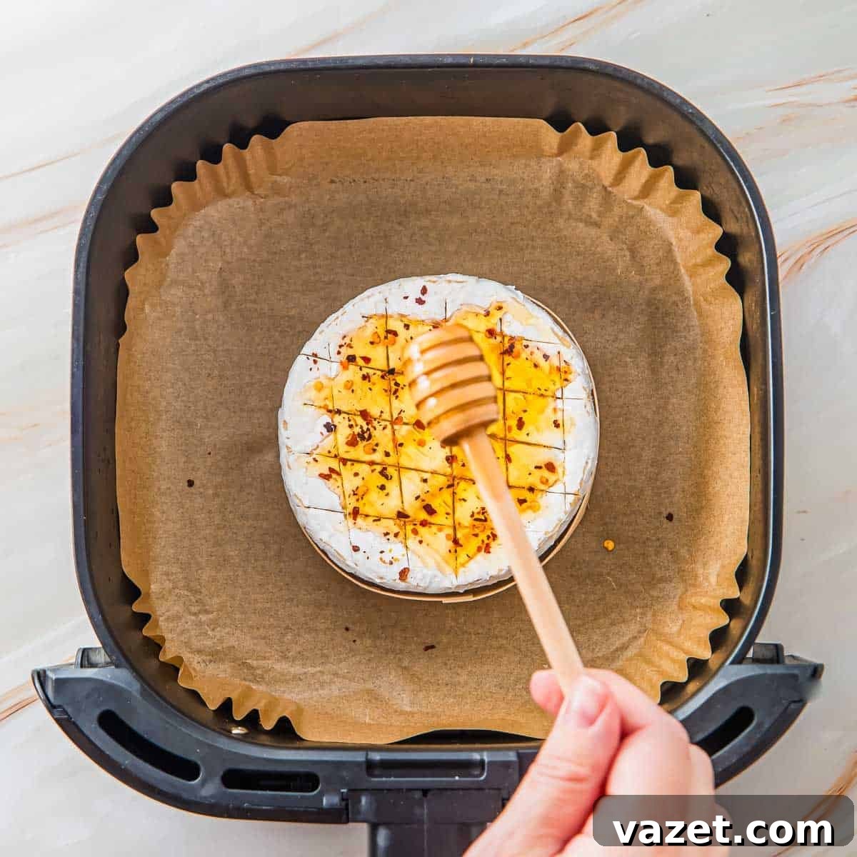 Honey-Kissed Baked Brie 5 A hand holds a honey dipper over a round wheel of brie cheese scored with a crosshatch pattern and sprinkled with chili flakes, placed on parchment paper inside an air fryer basket.