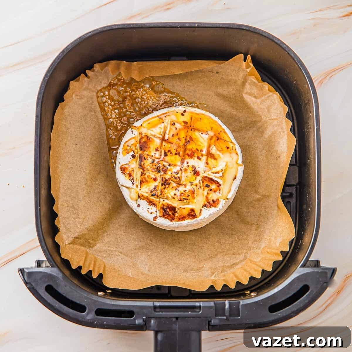 Honey-Kissed Baked Brie 6 A round wheel of baked brie cheese with a scored top sits on parchment paper inside an air fryer basket, melted cheese bubbling out on one side.