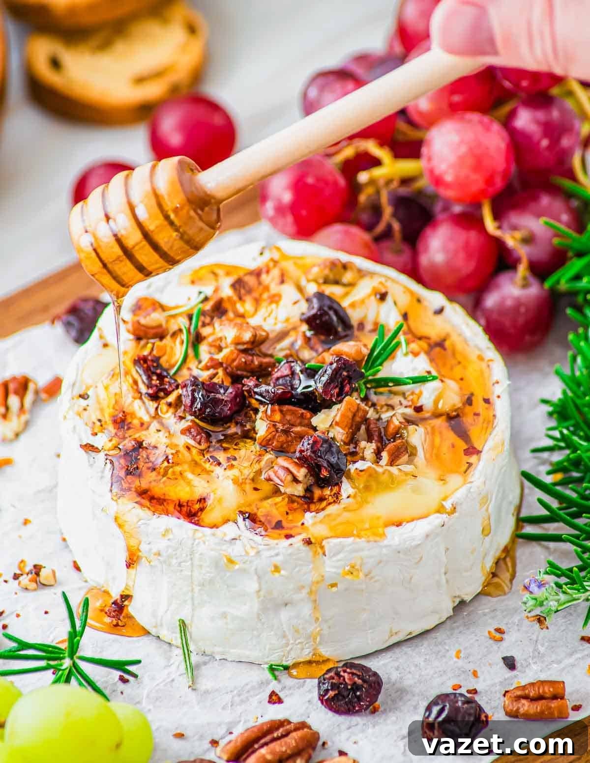 Honey-Kissed Baked Brie 7 A wheel of baked brie topped with honey, chopped pecans, dried cranberries, and rosemary, being drizzled with more honey. Red grapes, green grapes, rosemary sprigs, and crackers surround the cheese on a parchment-lined board.