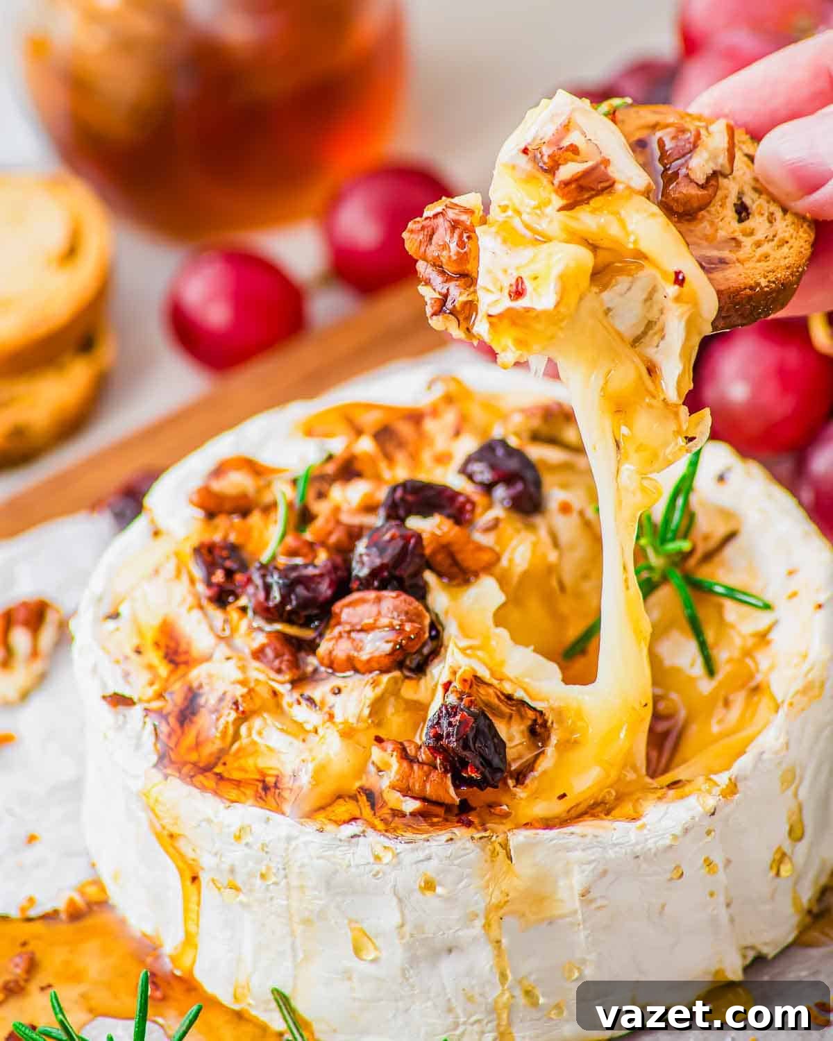 Honey-Kissed Baked Brie 9 A hand dips a toasted baguette slice into warm, gooey baked brie topped with honey, nuts, and dried cranberries. Fresh rosemary and grapes are seen nearby on the serving board.