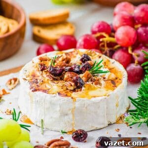 Honey-Kissed Baked Brie 10 A wheel of honey baked brie topped with chopped pecans, dried cranberries and rosemary sits on parchment paper, surrounded by green and red grapes, crackers, and fresh rosemary.