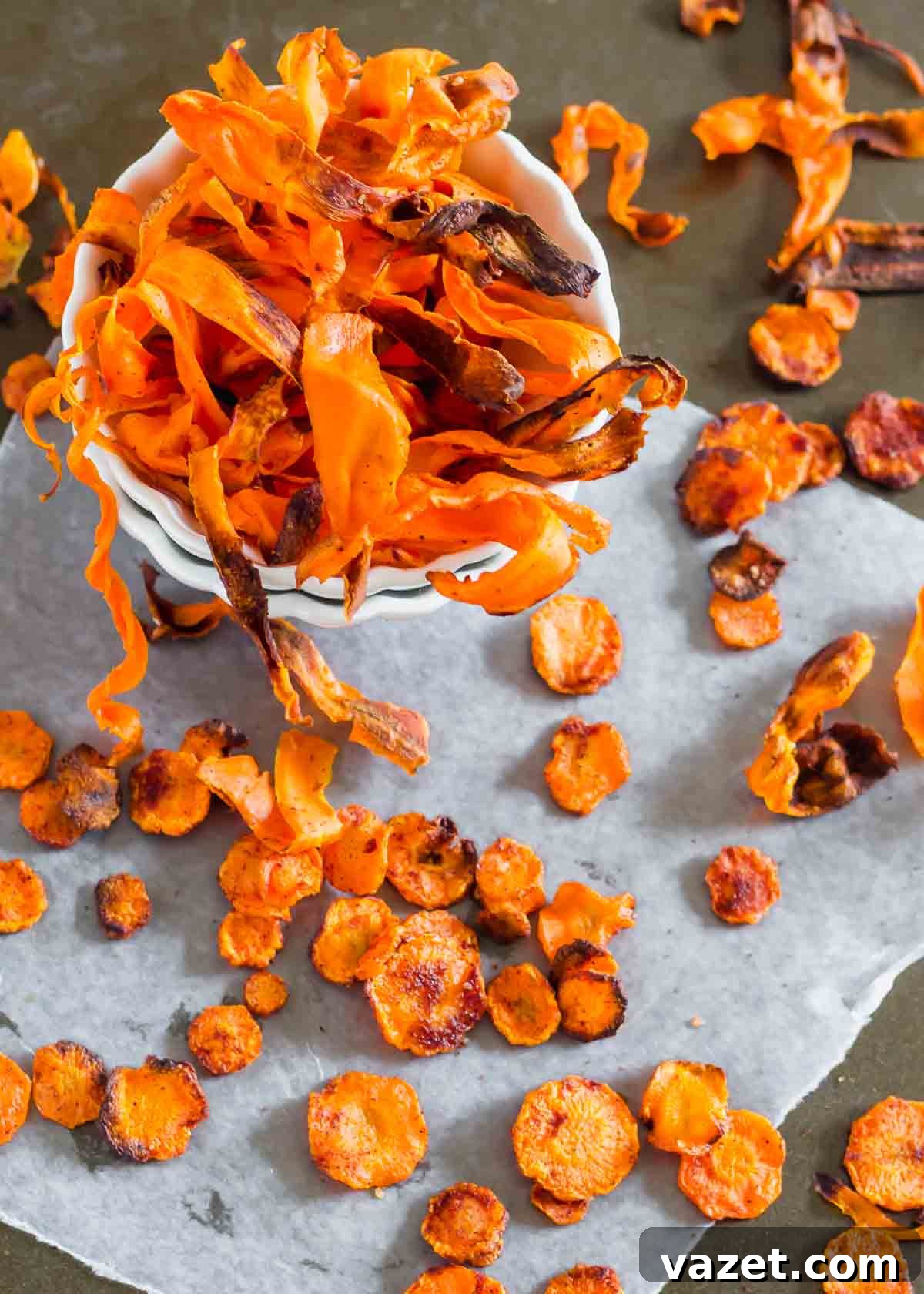Crunchy Roasted Carrot Crisps 4 A bowl of crispy carrot chips atop a slate surface surrounded by additional scattered chips, showing their vibrant color.