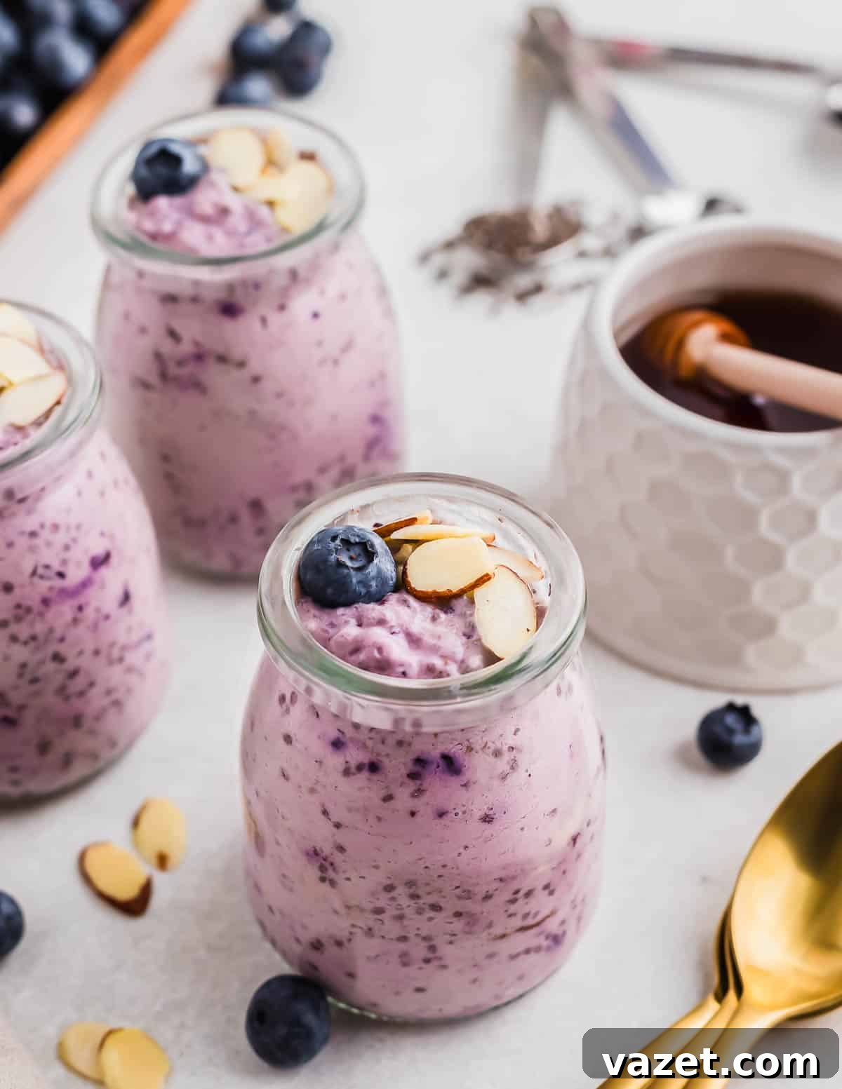 Berry Burst Chia Delight 3 Three small glass jars filled with blueberry chia pudding, topped with almond slices and blueberries, are placed in the foreground. A honey jar, spoon, and other ingredients are in the background.